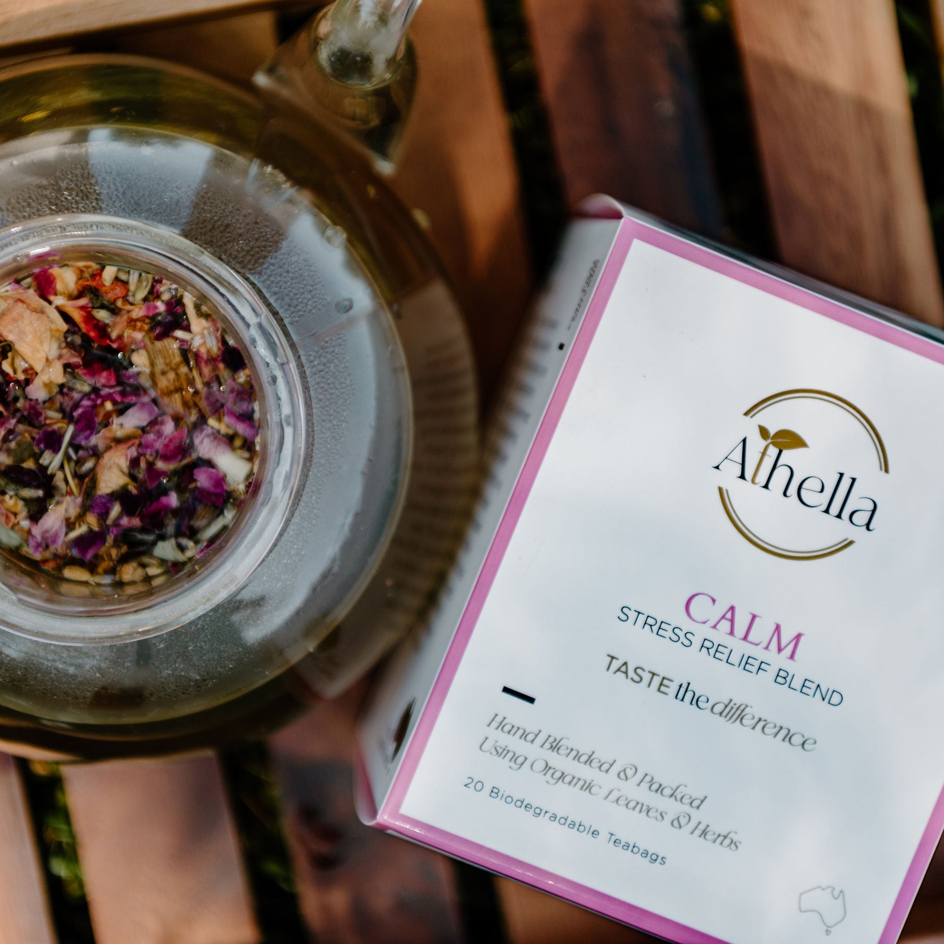 Athella Clam Stress Relief Blend box packaging next to clear teapot with loose leaf Calm tea leaves. Stress Relief Blend, Taste the Difference, Hand Blended and Packed Using Organic Leaves and Herbs. 20 Biodegradable Teabags.