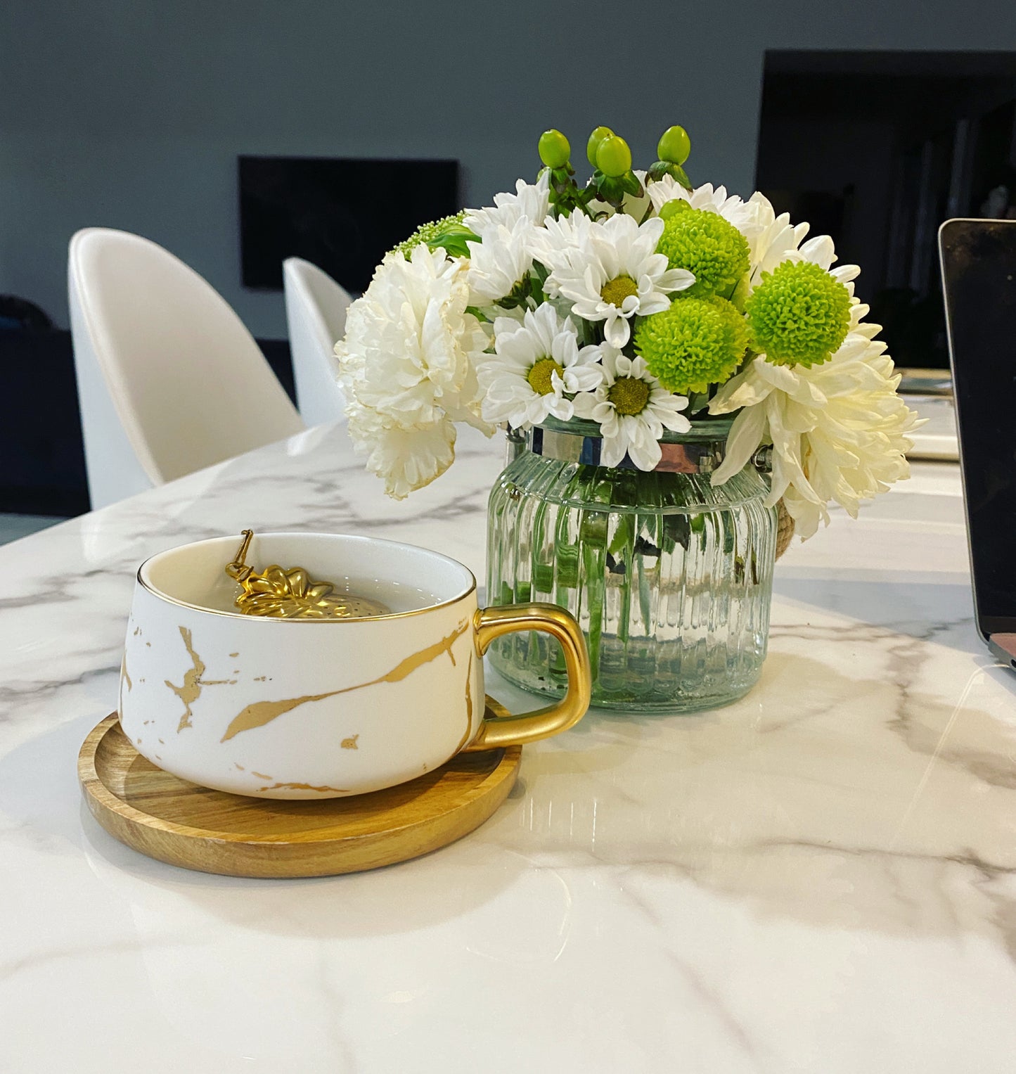 Athella Boho Luxe White and Gold Tea Cup on a Wooden Coaster with a Pineapple Tea Strainer styled on a marble bench with white flowers.